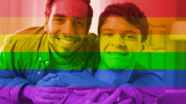 Портрет світлого чоловіка Gay Couple at Home. Людина сидить на дивані, його партнер витягує його з-за гола. Вони щасливі і посміхаються. Приміщення має сучасний інтер'єр. На ньому є фільтр прапора Свободи.. — стокове фото