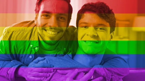 Retrato de una dulce pareja gay masculina en casa. Man Sits on a Couch, His Partner Embraces him from Behind. Son felices y sonrientes. Habitación tiene interior moderno. Shot tiene filtro Freedom Rainbow Flag en él. — Foto de Stock