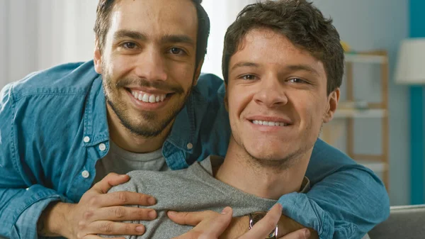 Portrait of a Gentle Male Gay Couple at Home. Young Man Sits on a Couch, His Partner Embraces him from Behind. They are Happy and Smiling. Room Has Modern Interior. — Stock Photo, Image