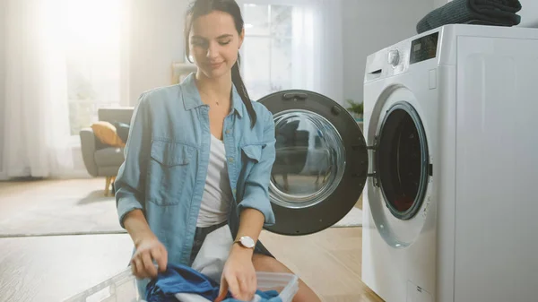 Belle brune souriante jeune femme assise devant un lave-linge en jeans Homely Clothes. Elle charge la laveuse avec Dirty Laundry. Salon lumineux et spacieux avec intérieur moderne. — Photo