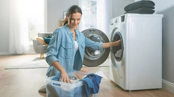Belle et heureuse jeune femme brune vient vers la machine à laver dans les vêtements Jeans Homely. Elle charge la laveuse avec Dirty Laundry. Salon lumineux et spacieux avec intérieur moderne. — Photo