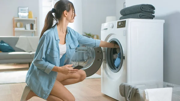 Belle jeune femme s'assoit à genoux à côté de la machine à laver. Elle a chargé la laveuse avec Dirty Laundry et configuré la laveuse avec son smartphone. Tourné dans le salon avec intérieur moderne. — Photo