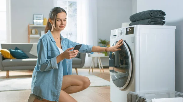 Belle jeune femme s'assoit à genoux à côté de la machine à laver. Elle a chargé la laveuse avec Dirty Laundry tout en utilisant son smartphone. Tourné dans le salon avec intérieur moderne. — Photo