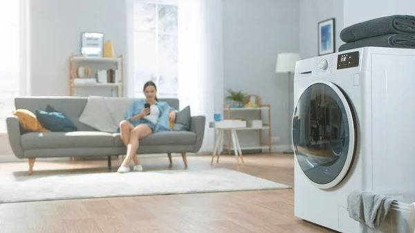 Tournage d'une machine à laver moderne technologiquement avancée travaillant dans un salon lumineux et spacieux avec un intérieur confortable. Jeune femme habillée à la maison utilise un téléphone intelligent en arrière-plan. — Photo