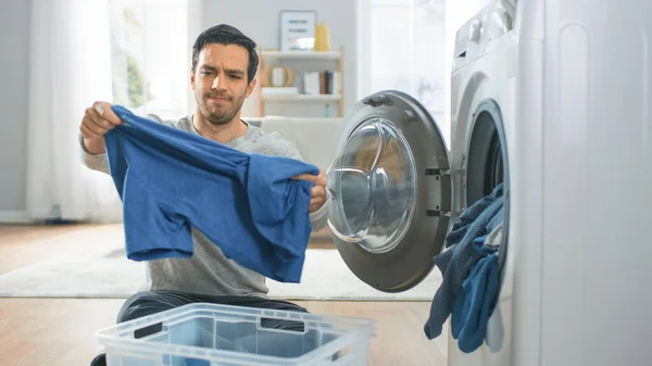 Bonito Jovem Confuso em Jeans Cinza e Casaco Senta-se na frente de uma máquina de lavar roupa em casa. Ele carrega a arruela com roupa suja. Sala de estar brilhante e espaçosa com interior moderno. — Fotografia de Stock