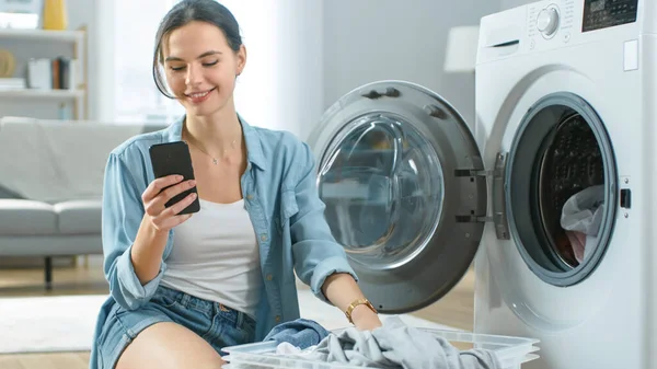 Mulher jovem sorridente bonita em roupas caseiras Senta-se na frente da máquina de lavar roupa e usa seu smartphone. Ela carrega a arruela com roupa suja. Sala de estar brilhante e espaçosa com interior moderno. — Fotografia de Stock