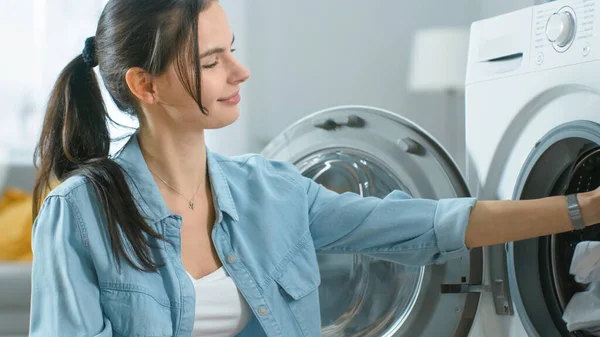 Gros plan d'une belle jeune femme vêtue de vêtements confortables assise devant un lave-linge. Elle charge la laveuse avec Dirty Laundry. Salon lumineux et spacieux avec intérieur moderne. — Photo