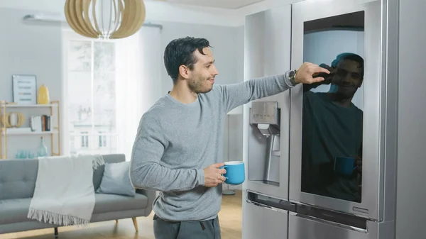 Όμορφος νεαρός άνδρας στέκεται δίπλα σε ένα ψυγείο, ενώ πίνοντας τον πρωινό καφέ του. Ελέγχει μια λίστα "To Do List" σε ένα Smart Fridge στο σπίτι. Κουζίνα είναι φωτεινό και άνετο. — Φωτογραφία Αρχείου