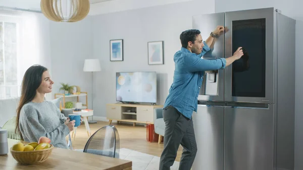 Hermosa pareja joven beber café de la mañana en la cocina. Hombre comprueba el pronóstico del tiempo y una lista de cosas que hacer en un refrigerador inteligente en casa. Apartamento es luminoso y acogedor. — Foto de Stock