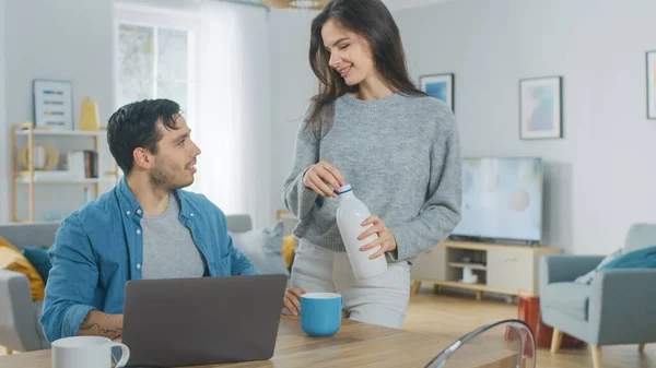 Beau jeune couple parlant dans une cuisine à la maison. L'homme travaille sur un ordinateur portable, Girl d verse du yaourt au lait laitier dans la tasse. Ils sont heureux et rient. Chambre a intérieur moderne. — Photo