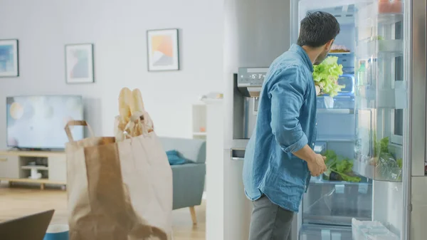 Bonito jovem atraente traz mantimentos para a cozinha. Ele coloca verdes salada fresca dentro da geladeira moderna. Quarto tem interior moderno brilhante. — Fotografia de Stock