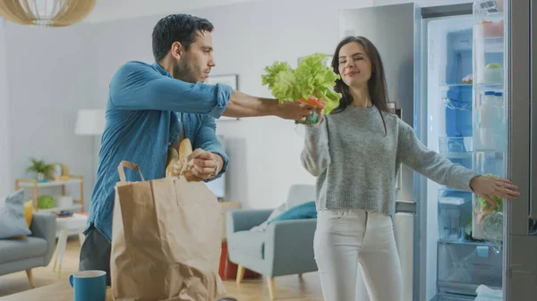 Beautiful Young Couple Come to the Kitchen with Fresh Fooceries in Brown Paper Bag. Man is Handing Fresh Salad Greens and Oranges to the Girl Who Puts them in the Fridge. — стоковое фото