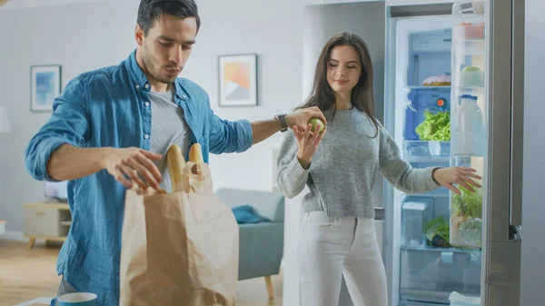 Beautiful Young Couple Come to the Kitchen with Fresh Fooceries in Brown Paper Bag. Человек протягивает свежие яблоки и апельсины девушке, которая кладет их в холодильник. — стоковое фото