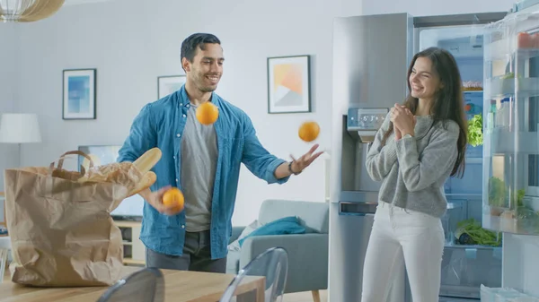 Krásný mladý pár Bavte se v kuchyni. Muž žongluje s pomeranči. Dívka tleská, povzbuzuje ho a smějí se. Pokoj má moderní Tech lednice s čerstvým potravinami. — Stock fotografie