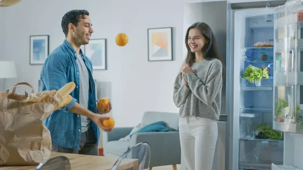 Krásný mladý pár Bavte se v kuchyni. Muž žongluje s pomeranči. Dívka tleská, povzbuzuje ho a smějí se. Pokoj má moderní Tech lednice s čerstvým potravinami. — Stock fotografie