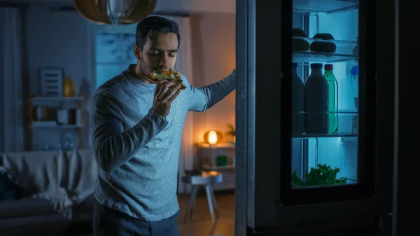 Guapo atractivo joven está en la cocina por la noche. Está hambriento, Heladera está abierta y muerde un pedazo de pizza sobrante. Lo está comiendo y se siente satisfecho. —  Fotos de Stock