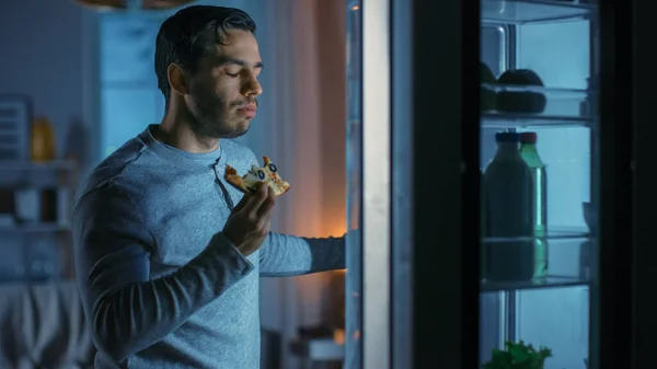 At Night in the Kitchen Handsome Attractive Young Man is Eating a Leftover Pizza from the Fridge. He is Hungry and Feels Satisfied.