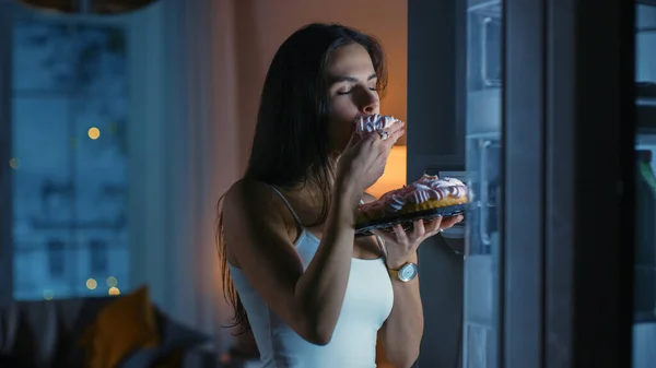 En la cocina en la noche hambrienta hermosa mujer joven muerde un pedazo de un delicioso pastel cremoso. Ella se siente satisfecha. — Foto de Stock