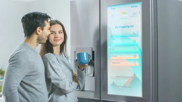 Hermosa pareja joven beber café de la mañana en la cocina. Verifican el pronóstico del tiempo y una lista de cosas que hacer en un refrigerador inteligente en casa. Apartamento es luminoso y acogedor. — Foto de Stock