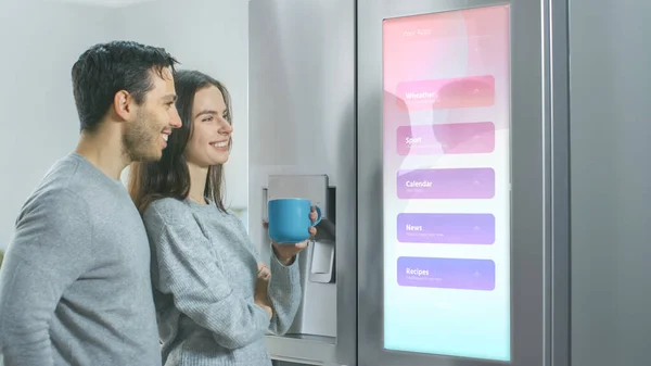 Hermosa pareja joven beber café de la mañana en la cocina. Verifican el pronóstico del tiempo y una lista de cosas que hacer en un refrigerador inteligente en casa. Apartamento es luminoso y acogedor. — Foto de Stock