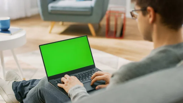 Over the Shoulder Shot: Young Man Wearing Glasses Funciona em um Computador Laptop com Tela Mock-up Verde. Ele está sentado em um sofá em sua acolhedora sala de estar. — Fotografia de Stock