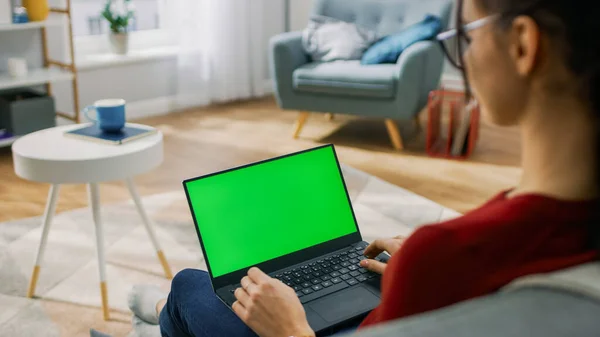 Молода жінка вдома працює на портативному комп'ютері з зеленим макетним екраном. Сидить на дивані у своїй затишній вітальні. Над плечовим пострілом — стокове фото