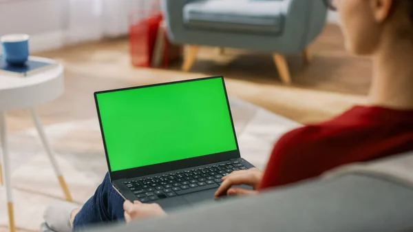 Young Woman at Home Works on a Laptop Computer with Green Mock-up Screen. Se sienta en un sofá en su acogedora sala de estar. Sobre el hombro disparado —  Fotos de Stock
