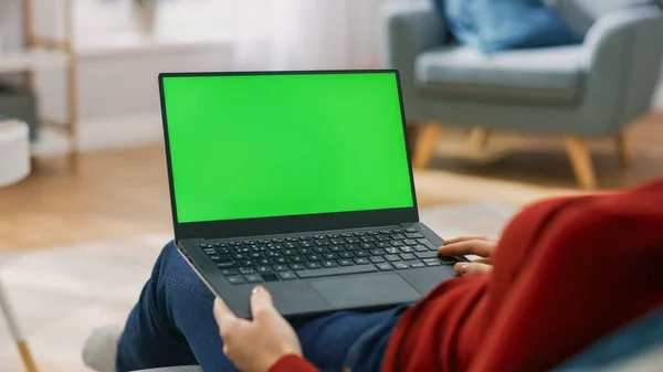 Jovem mulher em casa trabalha em um computador portátil com tela de falsificação verde. Ela está sentada em um sofá em sua acolhedora sala de estar. Sobre o tiro no ombro — Fotografia de Stock