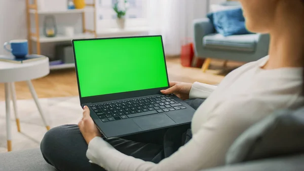 Mujer joven en casa sentada en un sofá trabaja en una computadora portátil con pantalla verde simulada. Chica Usando Computadora, Navegando por Internet, Ver Contenido, Chatear en Redes Sociales con Amigos. —  Fotos de Stock