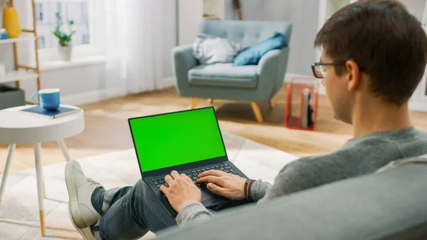 Ο νεαρός άνδρας στο σπίτι εργάζεται σε έναν φορητό υπολογιστή με πράσινη οθόνη mock-up. Κάθεται σε έναν καναπέ στο άνετο σαλόνι του. Πάνω από τον ώμο Shot. — Φωτογραφία Αρχείου