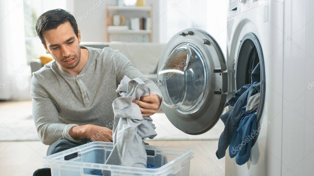 Handsome Smiling Young Man in Grey Jeans and Jumper Sits in Front of a Washing Machine at Home. He Loads the Washer with Dirty Laundry. Bright and Spacious Living Room with Modern Interior.