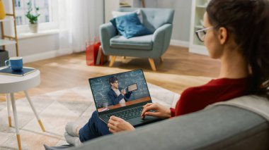 Evde Genç Bir Kadın Öğretmeniyle Video Görüşmesi İçin Dizüstü Bilgisayar Kullanıyor. Ekranda konuyu açıklayan öğretmenle çevrimiçi ders gösteriliyor. Oturma odasındaki kanepede oturuyordu. Omzunun üstünden.