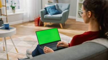 Green Mock-up ekranlı dizüstü bilgisayarda çalışan genç bir kadın. Oturma odasındaki bir koltukta oturuyordu. Omzunun üstünden.
