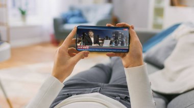 Genç Kadın Kanepede uzanıyor ve Yatay Peyzaj Modu 'nda Smartphone Haberleri' ni izliyor. Kız Cep Telefonu Kullanıyor, İçerik İzleme, Fren Haberleri. POV.