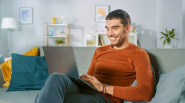 Yakışıklı adam evdeki koltukta otururken dizüstü bilgisayar kullanıyor. Adam çalışıyor, rahat oturma odasından interneti inceliyor..