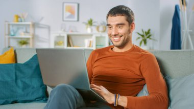 Yakışıklı adam evdeki koltukta otururken dizüstü bilgisayar kullanıyor. Adam çalışıyor, rahat oturma odasından interneti inceliyor..