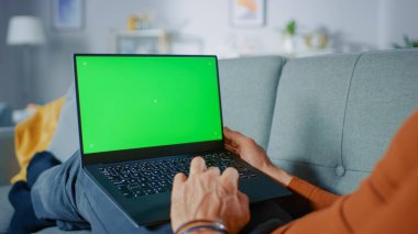 Evdeki kanepesinde yatan adam, Yeşil Model ekranlı dizüstü bilgisayar kullanıyor. Oturma odasında Anonim Adam 'ın Omuzdan Vuruşu.