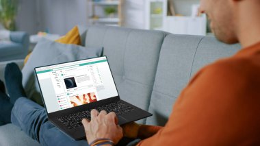 Yakışıklı Genç Adam Evde Oturuyor Laptop kullanıyor ve Sosyal Ağ Besleme Sitesi 'nde kürek çekiyor. Oturma odasında Anonim Adam 'ın Omuzdan Vuruşu.