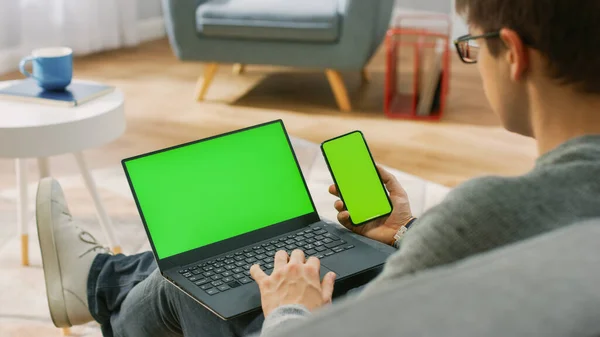 Young Man at Home arbeitet an einem Laptop mit grünem Bildschirm, während er ein Smartphone mit Chroma-Schlüsselanzeige in der Hand hält. Er sitzt auf einer Couch in seinem gemütlichen Wohnzimmer. Über die Schulter geschossen — Stockfoto