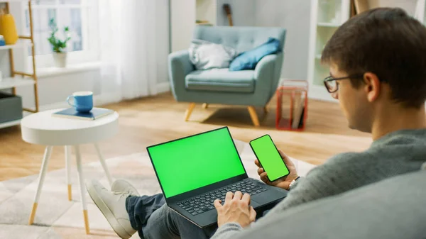 Young Man at Home Funciona em um Computador Laptop com Tela Mock-up Verde, enquanto segura Smartphone com Chroma Key Display. Ele está sentado em um sofá em sua acolhedora sala de estar. Sobre o tiro da câmera do ombro — Fotografia de Stock