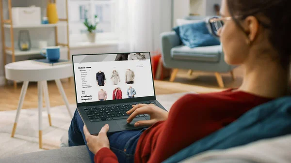 Jeune femme à la maison à l'aide d'un ordinateur portable pour naviguer sur le site de vente au détail en ligne. Elle est assise sur un canapé dans son confortable salon. Par-dessus l'épaule caméra tir — Photo