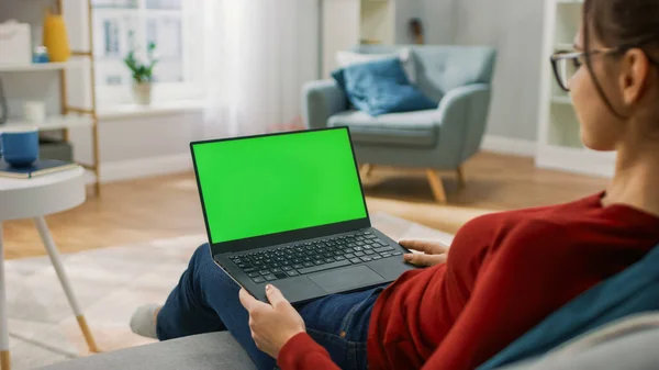 自宅の若い女性は、緑のモックアップ画面とノートパソコンのコンピュータ上で動作します.彼女は彼の居心地の良いリビングルームでソファに座っています.肩のショットの上に — ストック写真