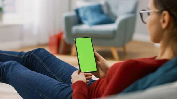 在家里的年轻女人使用绿色模拟屏幕智能手机。她坐在舒适的客厅里的长椅上。越过肩膀开枪了 — 图库照片
