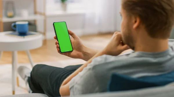 Young Man at Home utilizza lo smartphone con schermo finto verde. E 'seduta su un divano nel suo accogliente salotto. Sopra la spalla Colpo — Foto Stock
