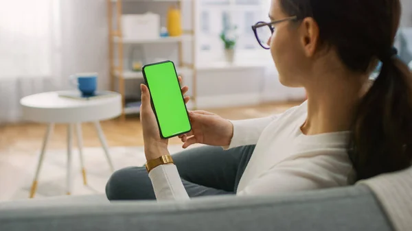 Jeune femme à la maison utilise un smartphone à écran vert maquillé. Elle est assise sur un canapé dans son confortable salon. Par-dessus l'épaule — Photo