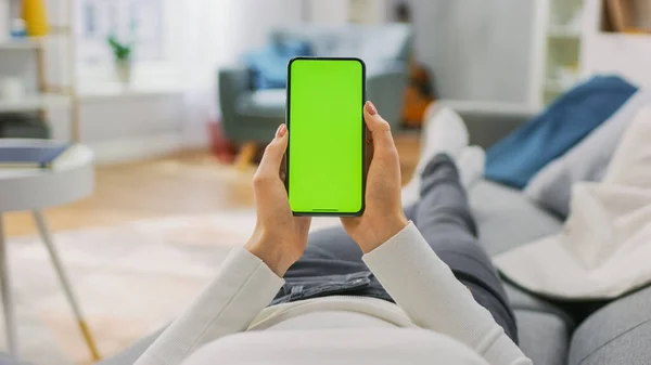 Giovane donna a casa utilizza Green Mock-up Screen Smartphone. E 'seduta su un divano nel suo accogliente salotto. Punto di vista girato. — Foto Stock