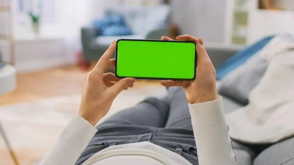 Jeune femme à la maison posée sur un canapé à l'aide d'un téléphone intelligent Green Mock-up Screen en mode paysage horizontal. Fille en utilisant le téléphone mobile, Naviguer sur Internet, Regarder du contenu, Vidéos, Blogs. Point de vue. — Photo