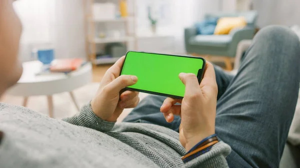 Man at Home Sdraiato su un divano con Smartphone, lo tiene orizzontalmente in modalità paesaggio. Premere i pulsanti per giocare al videogioco. Punto di vista Fotocamera girato. — Foto Stock