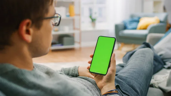 Man at Home Piesting on a Couch segítségével Smartphone zöld Mock-up Screen. Guy használata mobiltelefon, Internet Social Networks Böngészés. — Stock Fotó