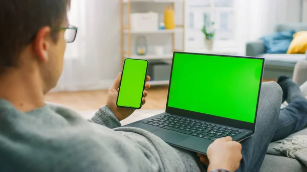 Young Man at Home Funciona em um Computador Laptop com Tela Mock-up Verde, enquanto segura Smartphone com Chroma Key Display. Ele está sentado em um sofá em sua acolhedora sala de estar. Sobre o tiro no ombro — Fotografia de Stock